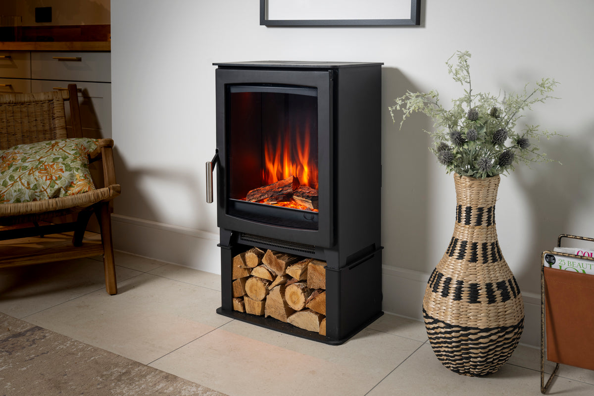 A black electric fireplace with a glass front door showing glowing orange flames, standing on a tiled floor next to a woven basket vase with dried flowers.