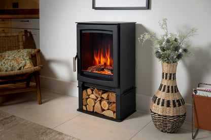 A black electric fireplace with a glass front door showing glowing orange flames, standing on a tiled floor next to a woven basket vase with dried flowers.