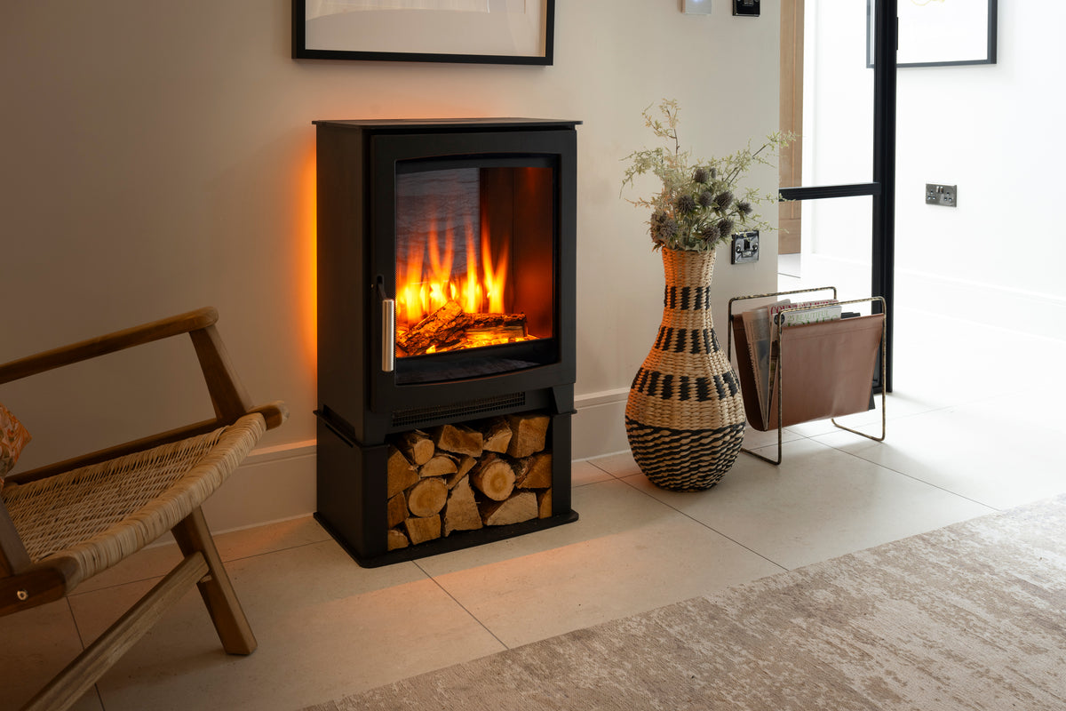 A black electric fireplace with a glowing fire inside, set in a modern living room next to a woven chair, a large patterned vase, and a magazine rack.