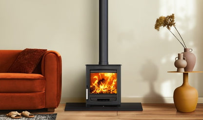 A black wood-burning stove with a visible fire, set in a cozy living room next to an orange armchair and a side table with vases.