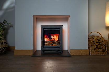 A black electric freestanding fireplace insert with a glowing orange and yellow fire inside, set in a recessed wall opening with a light gray wall and dark wood floor. A basket of firewood and a lamp are visible on either side.