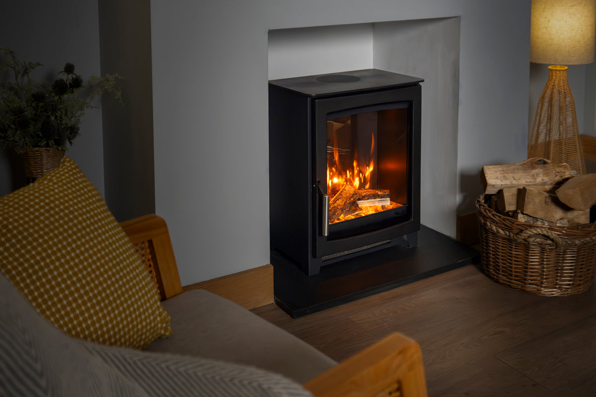 A black electric fireplace insert with a glowing fire inside, set in a modern room with a gray wall niche, wooden floor, a cozy armchair with a yellow patterned pillow, and a basket of firewood.