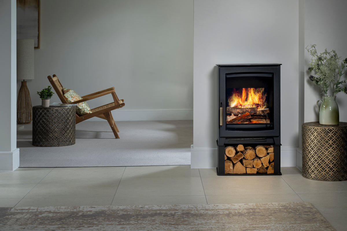 A black electric fireplace with a visible fire inside and stacked firewood in the lower compartment, shown in a styled interior room with a chair, side tables, and plants.