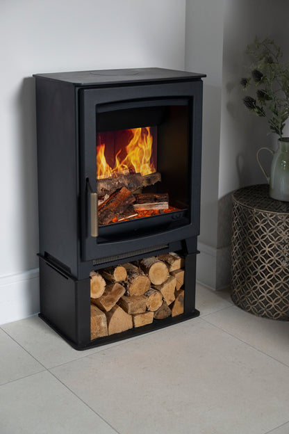 A black electric stove with a visible fire inside and a lower log storage compartment filled with cut firewood, shown in a real interior corner beside a woven side table and vase.