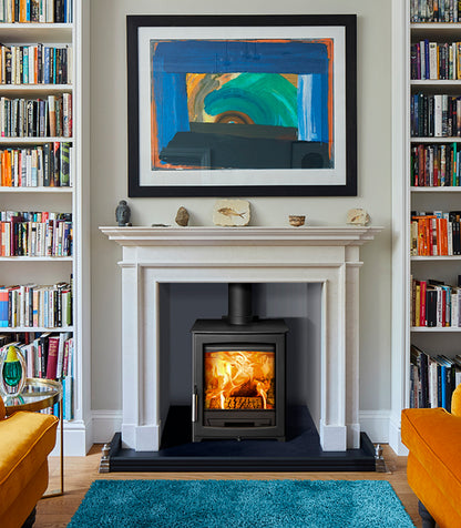 A black wood-burning stove with a glowing fire, set within a white fireplace mantel. The fireplace is styled in a cozy room with tall bookshelves on both sides, a blue abstract painting above, and a bright blue rug in the foreground.