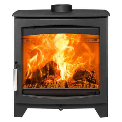 A black wood-burning stove with a glass front door showing a bright orange fire and logs inside, photographed on a plain white background. The brand name Puregran is visible on the lower right of the glass.
