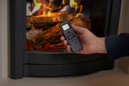 A hand holding a black remote control in front of a fireplace with logs and flames, demonstrating control of the fireplace.
