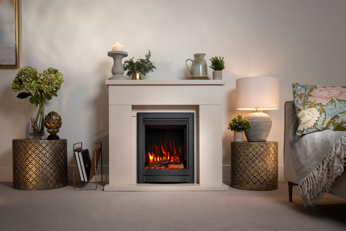 A modern electric fireplace insert with a black frame and glowing orange flames, set within a white stone mantel. The fireplace is styled in a cozy living room setting with a white side table, a lit table lamp, a patterned throw pillow, and decorative plants.