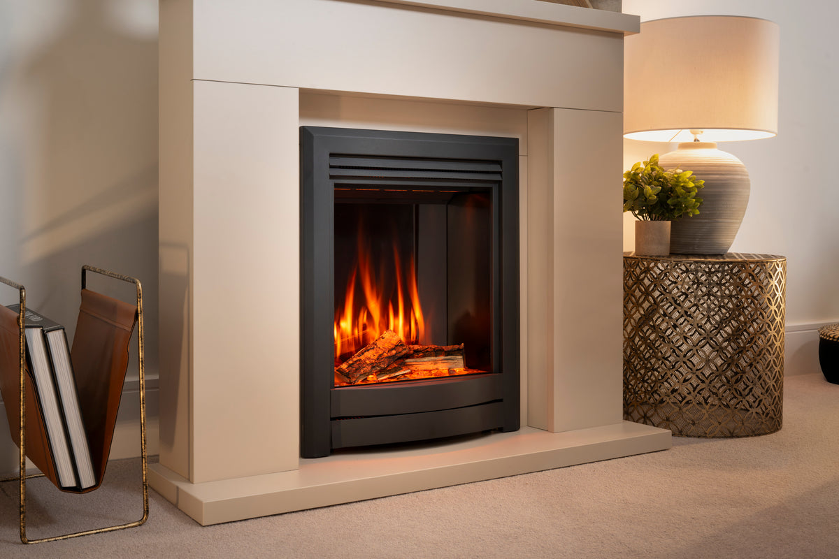 A modern black fireplace insert with a glowing orange and yellow fire, set within a cream-colored stone or concrete fireplace mantel. The scene is styled with a patterned side table holding a lamp and plant, and a magazine rack on the left.