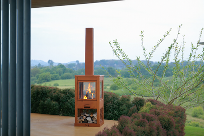 A freestanding metal wood-burning fireplace with a visible fire inside, set on a wooden decking surrounded by bushes in a landscaped garden overlooking fields and trees in the background.