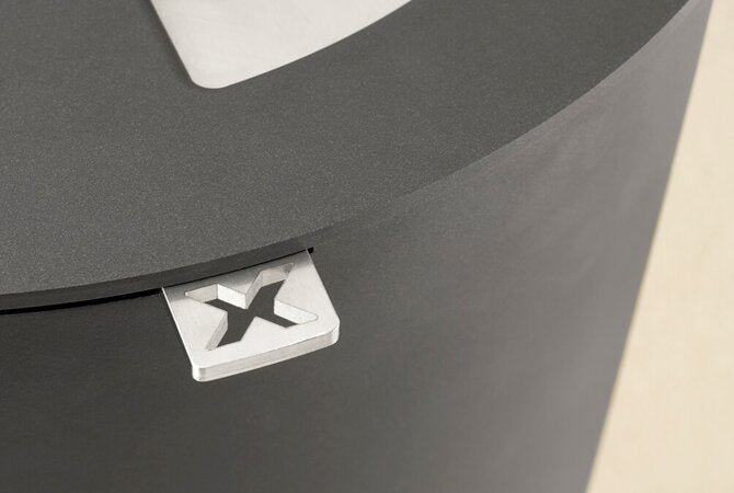 Close-up of a dark grey circular stove with a small white square detail featuring a black stylized letter X, on a light beige background.