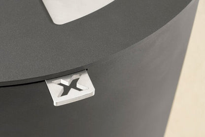Close-up of a dark grey circular stove with a small white square detail featuring a black stylized letter X, on a light beige background.