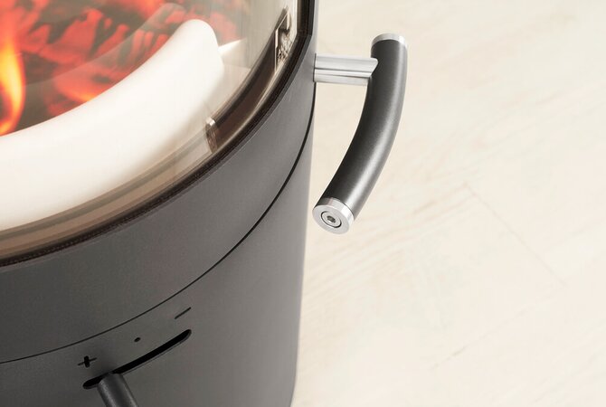 Close-up of a modern black cylindrical stove with a glass top showing glowing orange embers, a white inner liner, and a curved dark handle on a light wood floor.