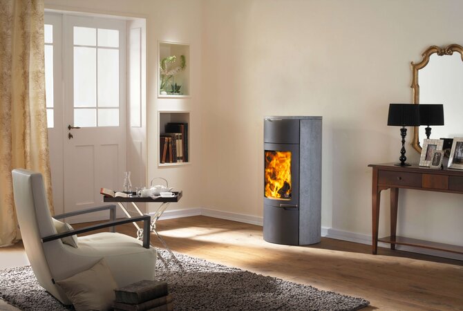 A modern gray wood-burning stove with a glowing fire, set in a cozy living room with a cream chair, wooden floor, and a console table with a mirror.