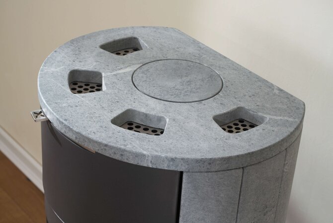 A close-up of a modern cylindrical appliance, likely a stove or heater, featuring a textured gray stone top with ventilation holes and a dark gray body, set against a plain beige wall.