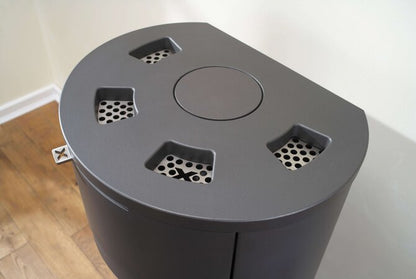 A dark gray metal cylindrical stove with slotted lid, photographed indoors against a plain wall and wood floor.