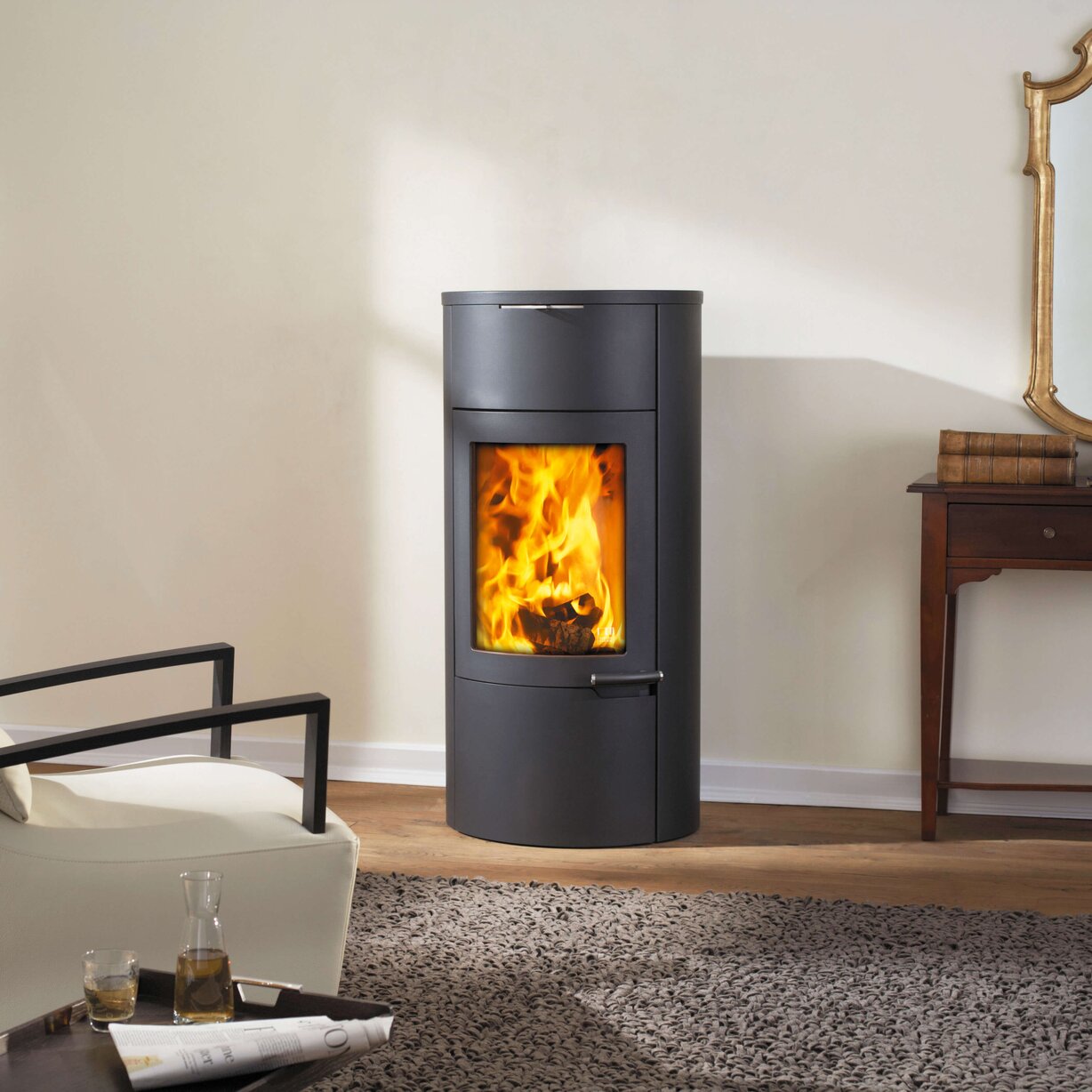 A modern dark gray cylindrical wood-burning stove with a glowing fire visible in the front window, set in a styled living room with a chair, side table, rug, and wall.