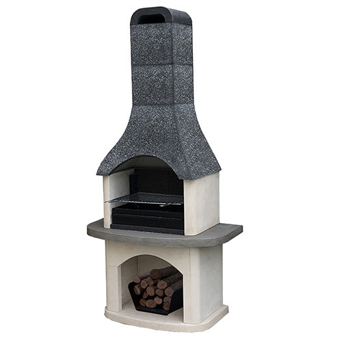 A freestanding outdoor stone barbecue grill with a dark textured chimney and light stone body, shown cooking food in a garden patio setting with plants, a wooden fence, and a dining table nearby.