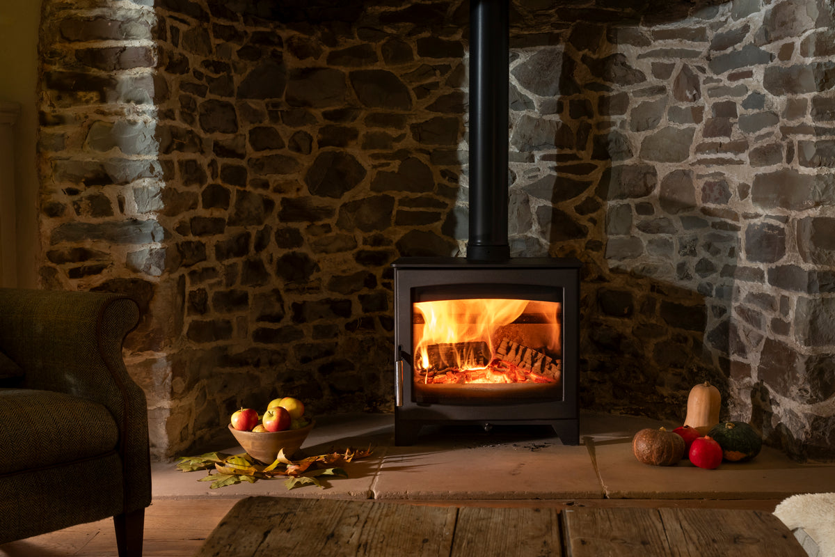 A black wood-burning stove with a glowing fire inside, set against a rustic stone fireplace wall. A dark upholstered armchair is visible on the left, and decorative pumpkins and apples sit on the hearth.