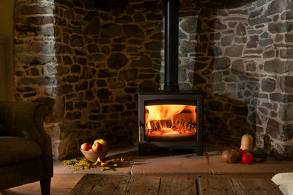 A black wood-burning stove with a glowing fire inside, set against a rustic stone fireplace wall. A dark upholstered armchair is visible on the left, and decorative pumpkins and apples sit on the hearth.