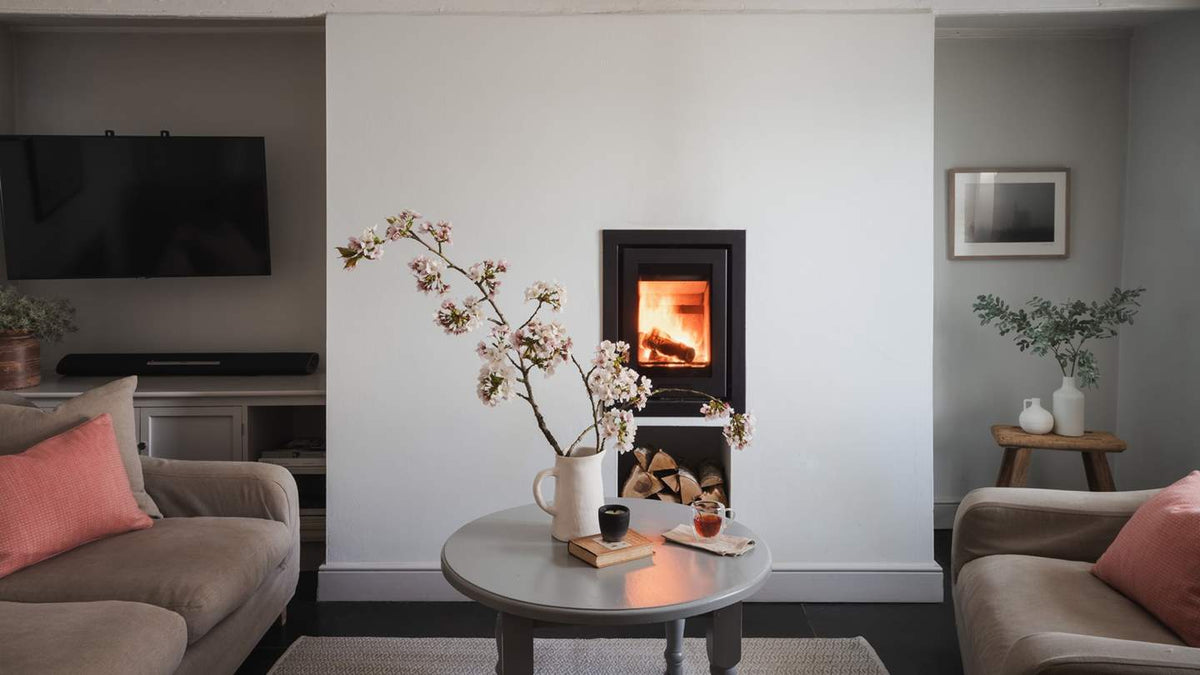 A modern black wood-burning fireplace insert set into a white wall in a styled living room with beige sofas, pink cushions, and a round coffee table in the foreground.