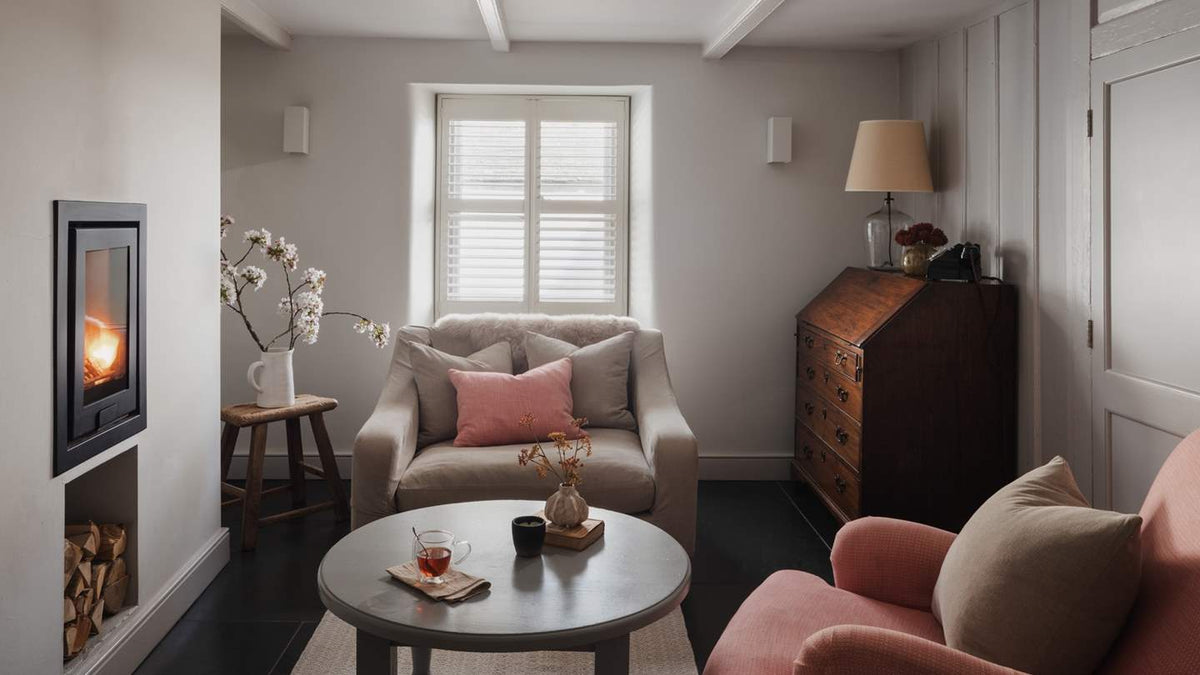A cozy living room interior featuring a beige armchair with pink and gray cushions, a round gray coffee table, a pink armchair in the foreground, a dark wood secretary cabinet, and a black fireplace insert with a glowing fire.