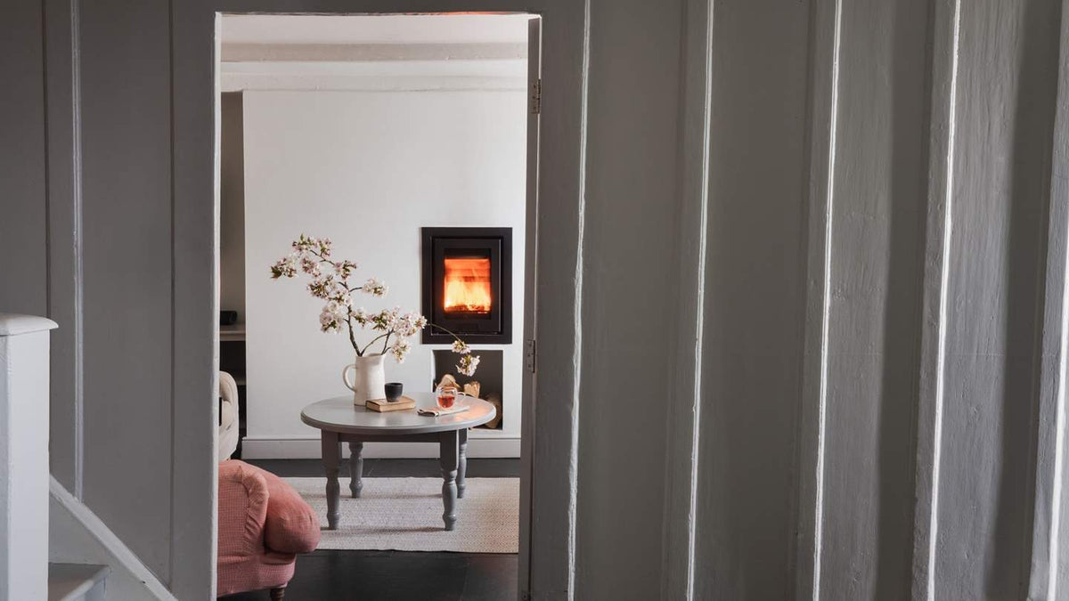 A view through a doorway into a cozy room with a round table, a vase of branches, and a black wood-burning fireplace. The foreground is framed by gray paneled doors.