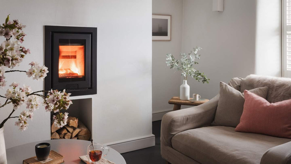 A modern black wood-burning fireplace with a glowing orange fire, set in a minimalist living room next to a beige sofa with pink cushions.