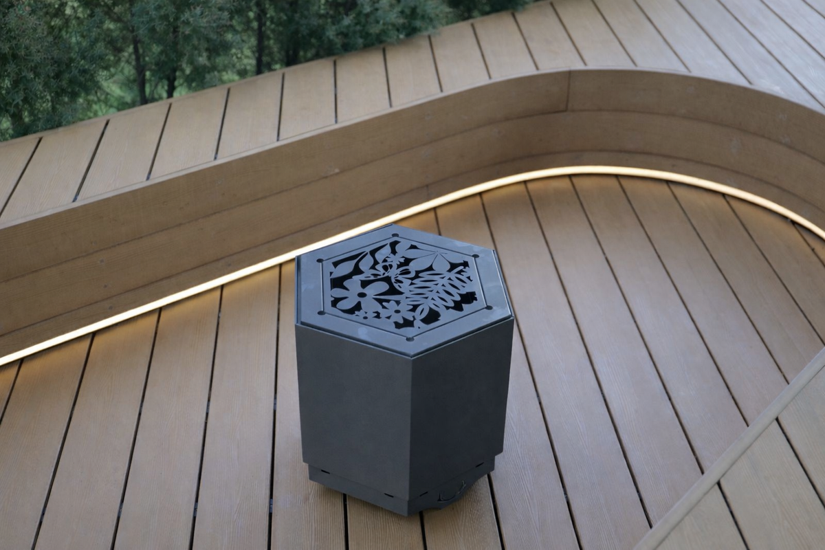 A dark grey hexagonal outdoor fire pit table with a decorative laser-cut floral pattern on top, set on a curved wooden deck with warm ambient lighting.
